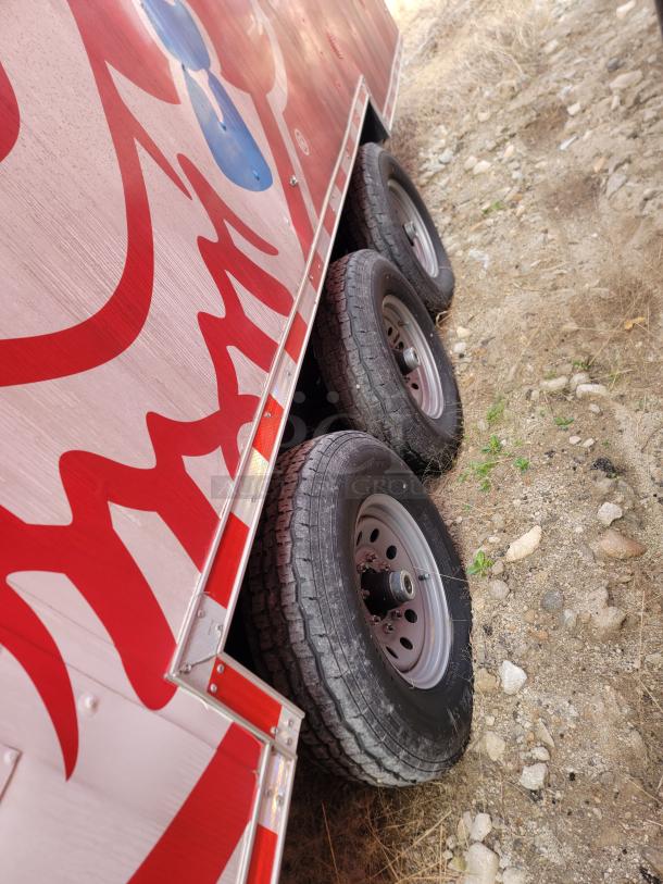 Side view of a red and white trailer with dual axle wheels, off-road. Visible wear on tires, red reflective strips on edges.