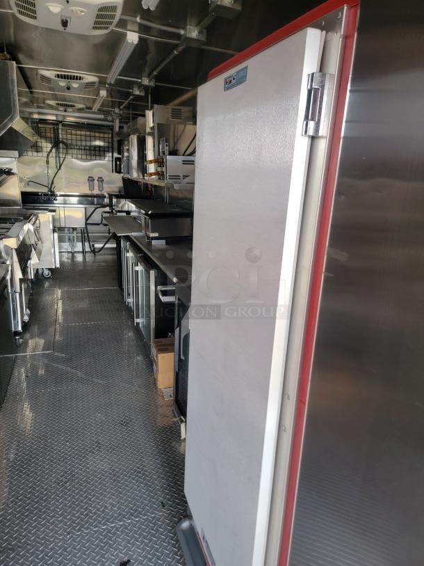 Commercial kitchen interior with stainless steel appliances, including a tall, silver refrigerator. Kitchen features metal flooring, overhead vents, and workstations.