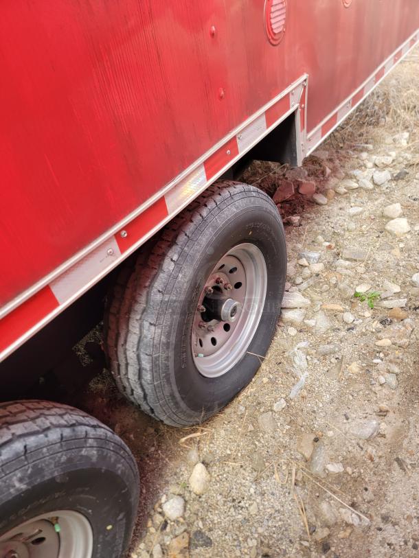 Red utility trailer, dual axle with visible wear on the tires. Reflective tape along lower edge. Located on rocky ground.