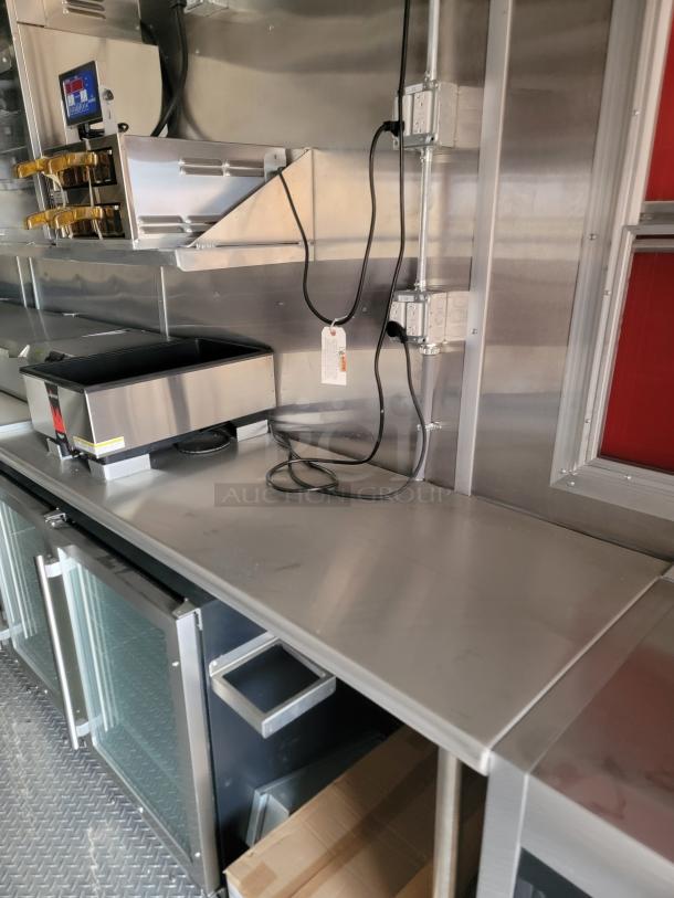 Stainless steel commercial kitchen workstation with mounted outlets, glass-door refrigerator, and equipment storage. Appears in excellent condition.