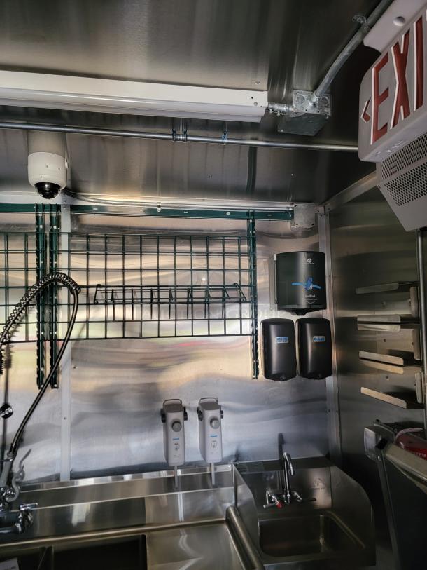 Industrial stainless steel kitchen with dual sinks, commercial sprayer faucet, wall-mounted soap dispensers, paper towel holders, and exit sign.