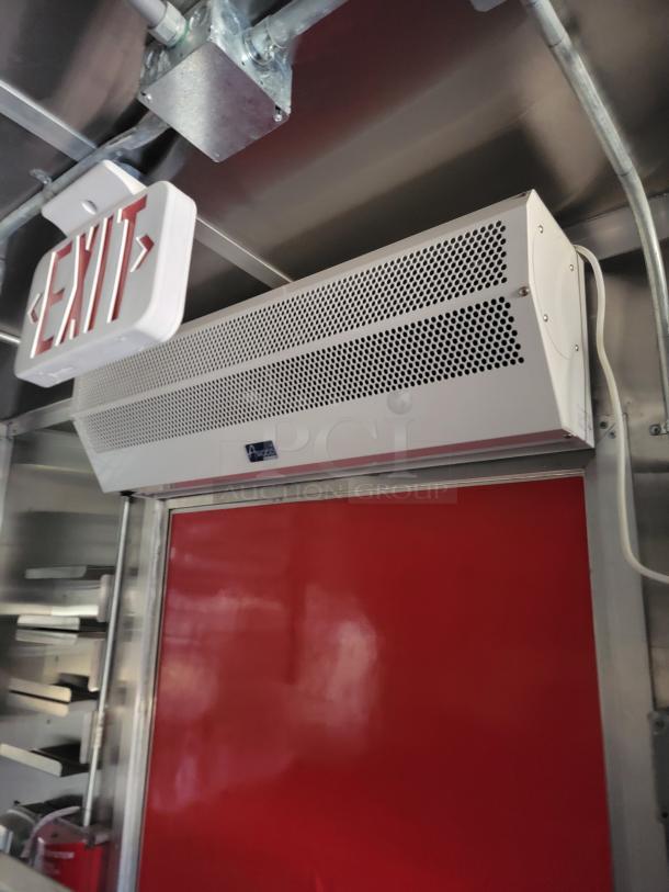 Ceiling-mounted air curtain above a red door, featuring a Vortex label. Nearby exit sign; metallic and wired setting.