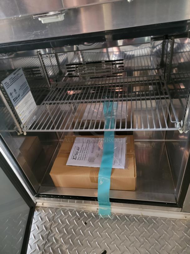 Turbo Air stainless steel refrigerator interior, featuring wire shelves and a cardboard box. Instruction sheet with warranty details visible.