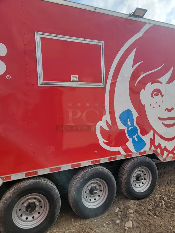 Red food truck trailer with a serving window, featuring a large graphic design. Triple axle wheels. Good condition.