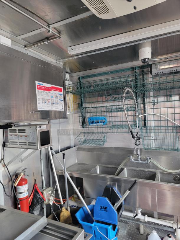 Stainless steel mobile kitchen interior with triple sink, overhead storage, Ecolab label, microwave, and cleaning supplies.