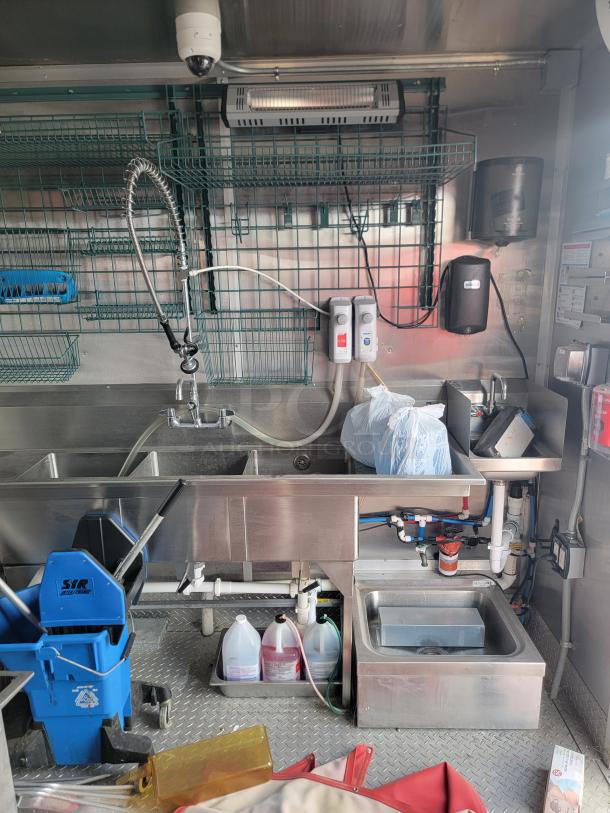 Commercial stainless steel kitchen setup with industrial sink, spray faucet, mixed plumbing system, and cleaning supplies.