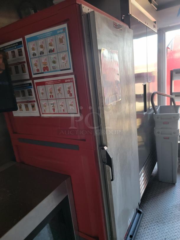 Commercial kitchen refrigerator with red panel, featuring instruction sheets on the door. Visible handle and metallic surface.