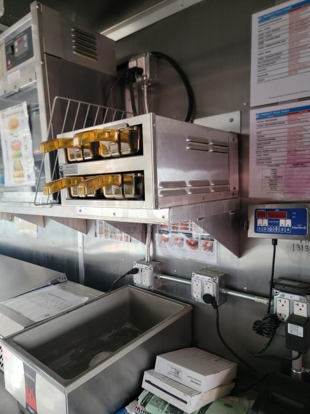 Commercial kitchen setup with stainless steel equipment, including dispensers with yellow handles, control panel, and outlet access.