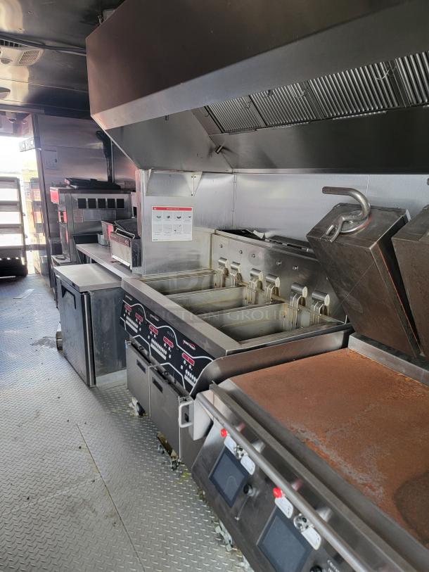 Commercial kitchen equipment including deep fryers, griddles, and a ventilation hood. Stainless steel finish, built-in storage.