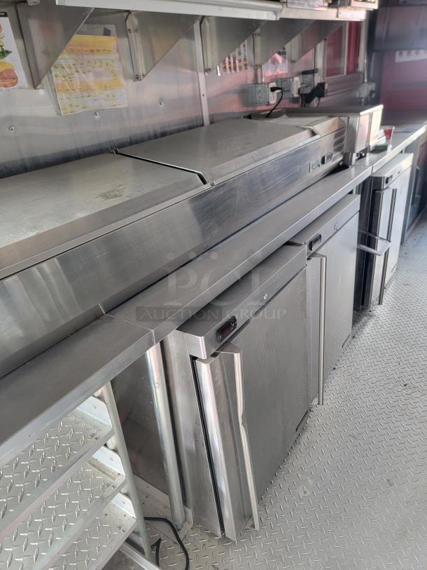 Stainless steel commercial kitchen counter with refrigeration units and prep area, in good condition with wall-mounted shelving.