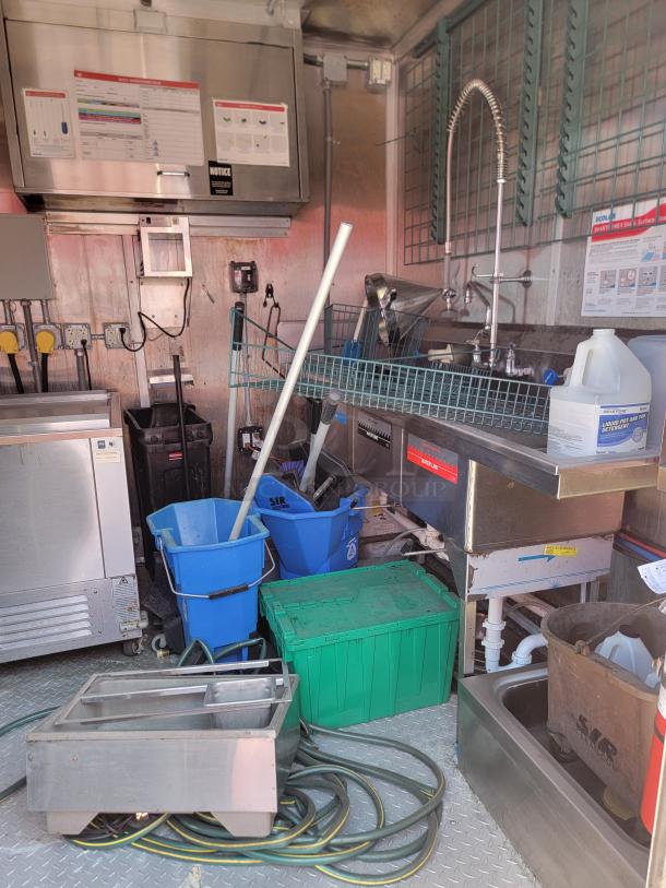 Commercial kitchen equipment setup with stainless steel sink, spray faucet, various cleaning supplies, buckets, and storage containers.