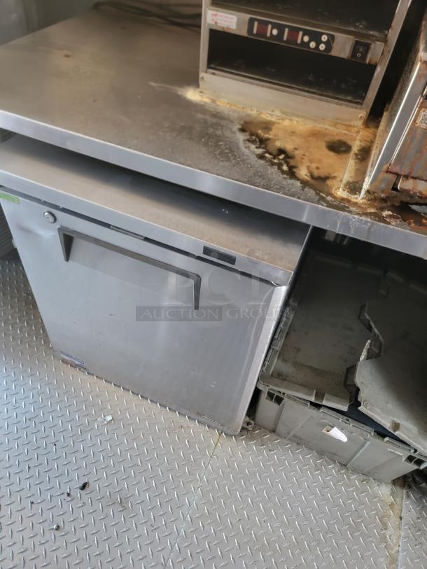 Commercial stainless steel under-counter refrigerator, showing signs of wear with rust and stains on top surface, near oven unit.