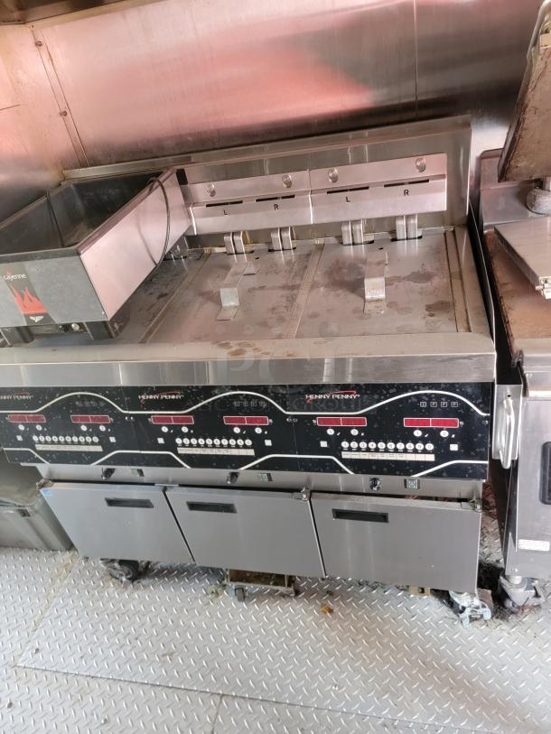 Henny Penny commercial fryer in stainless steel, shows signs of use. Features digital controls and several compartments.