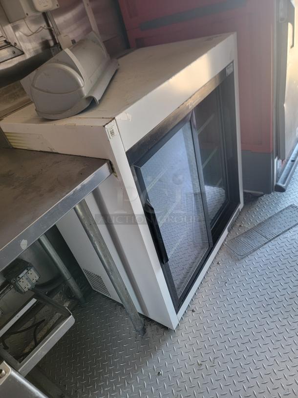 Commercial refrigerator with glass doors, white exterior, and visible shelving. Slight wear on the surface. Ideal for storage.