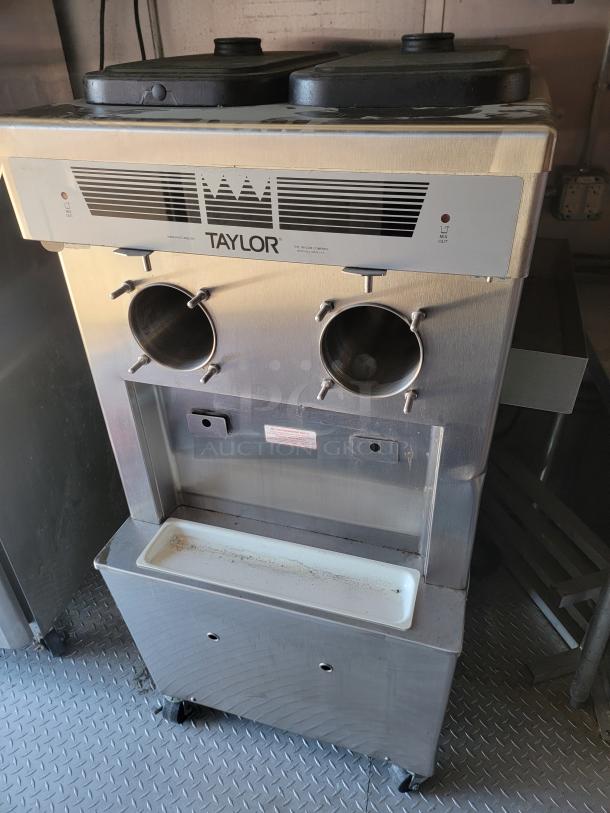 Taylor ice cream machine with dual dispensers, stainless steel, visible wear. Model and serial details on front panel.