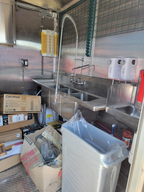 Commercial kitchen setup with stainless steel triple sink, industrial faucet, CyberPower equipment boxes, and waste bin.