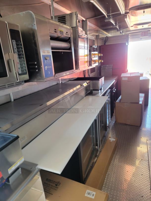 Food truck interior with stainless steel appliances, including a conveyor toaster, countertop griddle, and refrigerated display.