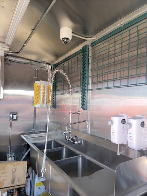 Commercial kitchen sink setup with stainless steel triple basin, industrial faucet, and white wall-mounted dispensers.