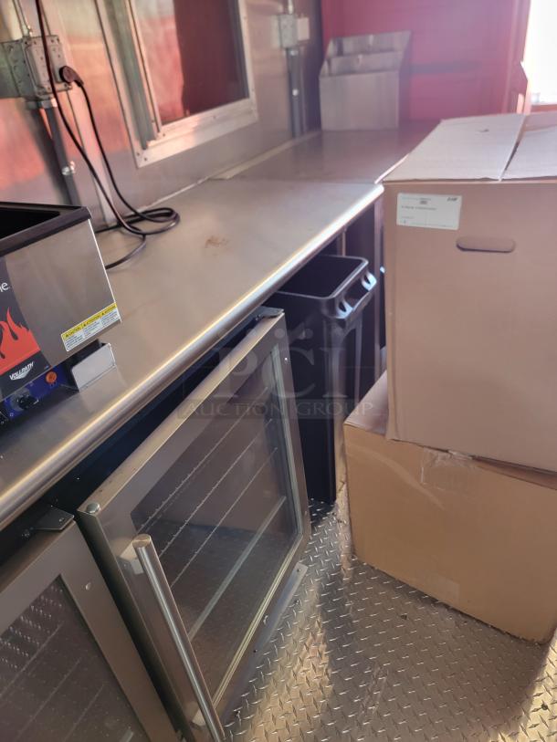 Stainless steel counter kitchenette with a dual glass-door fridge, cardboard boxes, and power cords visible. Clean condition.