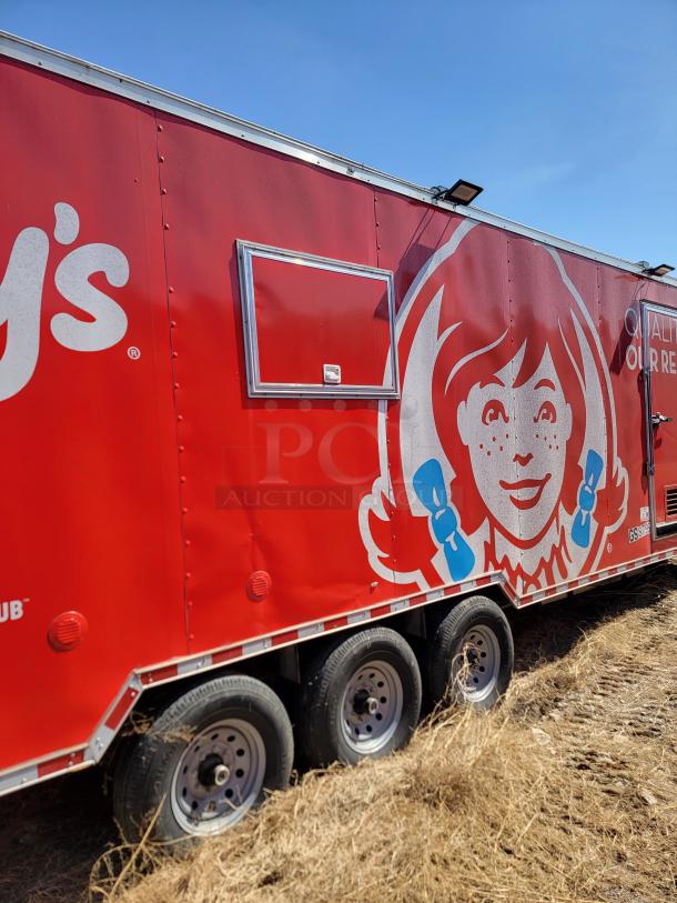 Red food truck trailer featuring Wendy's logo, showing three wheels and a serving window. Sturdy build, parked on dry grass.