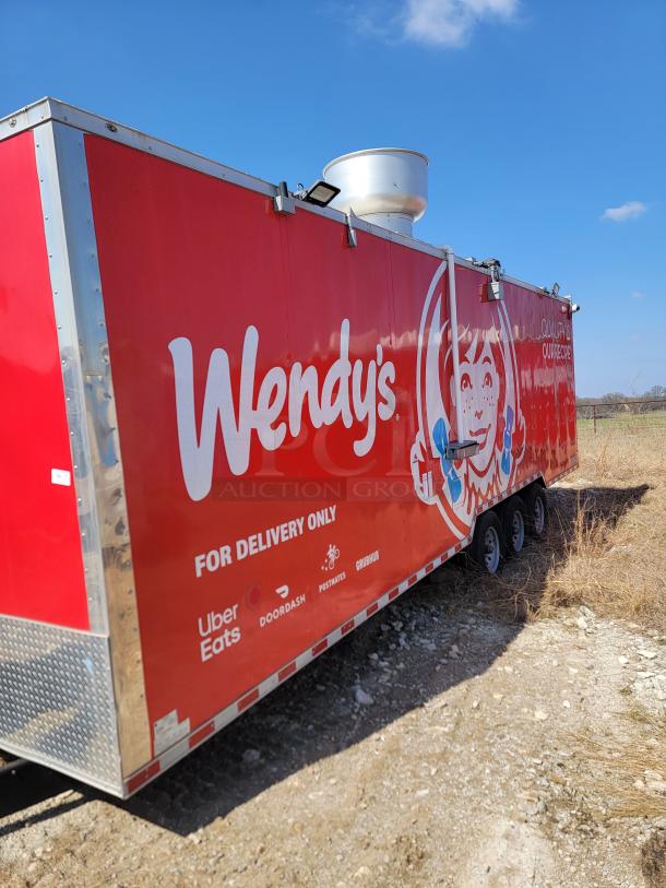 Red Wendy's food trailer with delivery service logos, triple-axle setup, and roof vent on a dirt lot under blue sky.