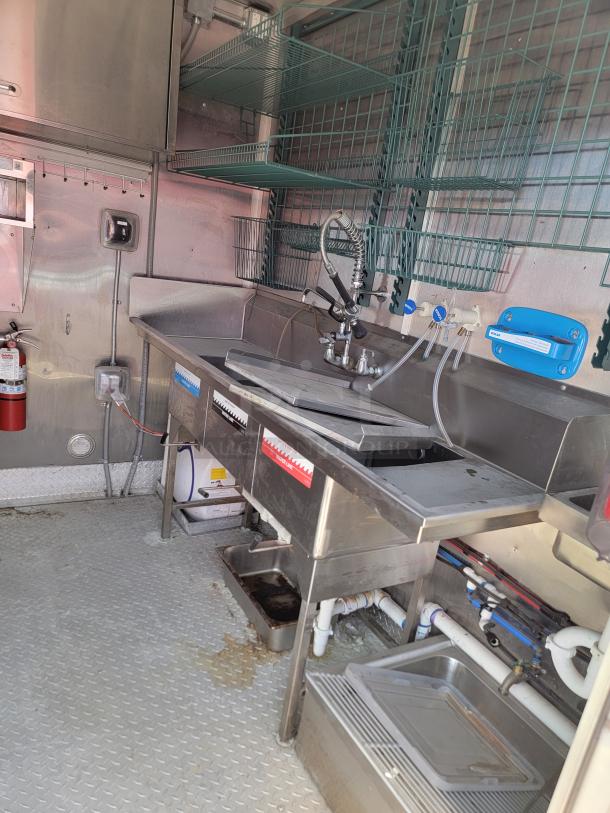Commercial stainless steel sink with sprayer, dual basins, and integrated shelves, in good condition. Wall-mounted utility connections.