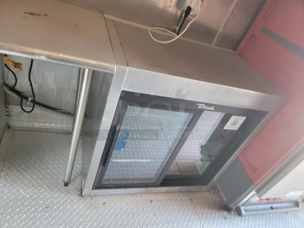 True glass door refrigerator, stainless steel top, dual sliding doors, electrical cable visible. Suitable for commercial use.