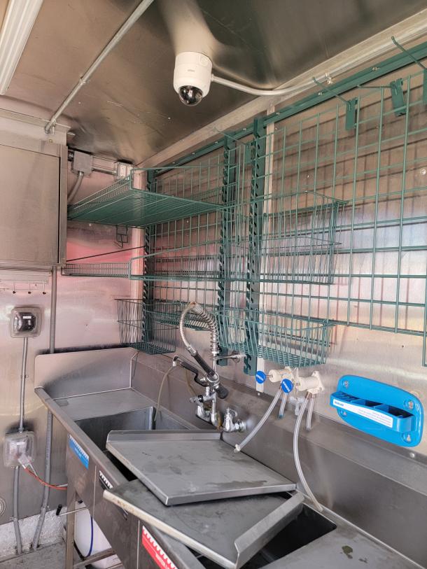 Commercial stainless steel sink with dual faucets, water filtering system, overhead security camera, and wire shelving units.
