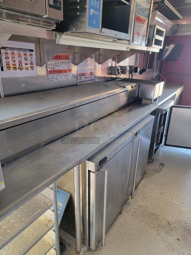 Stainless steel commercial prep counter with multiple storage units, slight wear visible. Menu posters and equipment on wall.