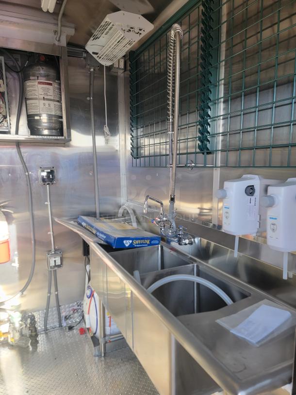 Commercial kitchen sink setup with three compartments, stainless steel construction, overhead sprayer, and various plumbing fixtures.