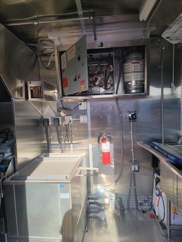 Industrial kitchen interior showcasing electrical panel with exposed wiring, stainless steel appliances, fire extinguisher, overhead piping, and equipment.