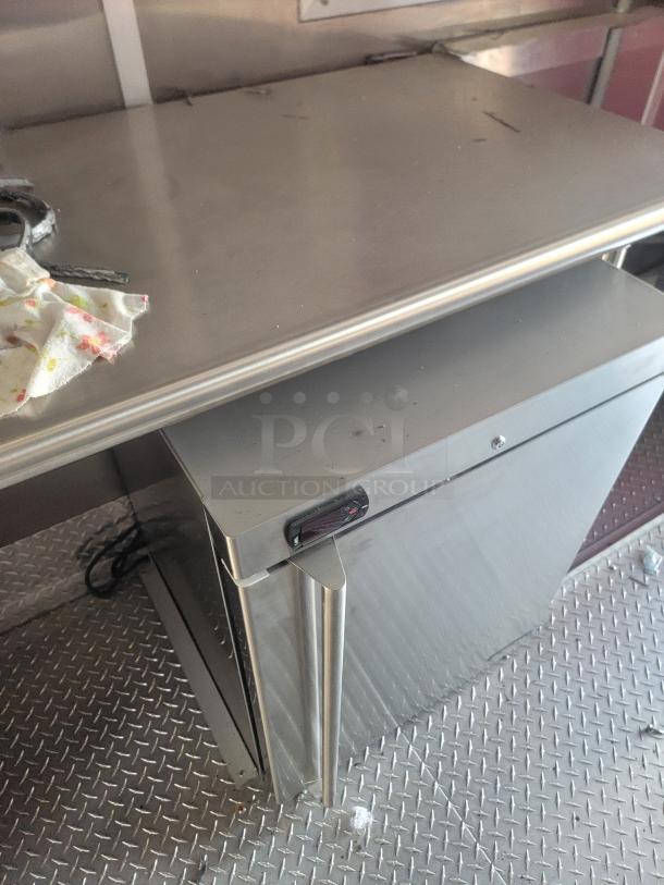 Stainless steel commercial refrigerator with digital control panel, located under a metal prep table, on a textured floor.