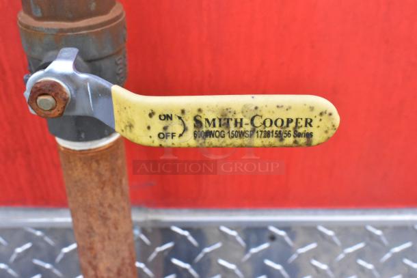 Close-up of a Smith-Cooper valve handle with "On/Off" markings, attached to a rusted pipe in front of a red background.