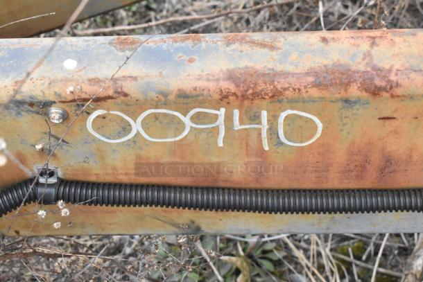 Rusty metal surface with the number "00940" written in white on it, surrounded by dry vegetation.