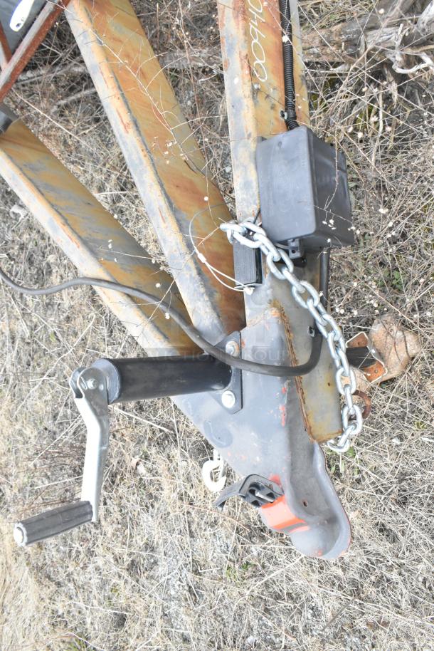 Trailer hitch with a chain and crank handle, rusted metal frame on dry grass. No equipment visible.