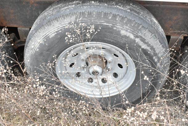 Trailer tire with steel rim, surrounded by dry grass. Tire shows some dirt and wear. Part of triple axle mobile kitchen.