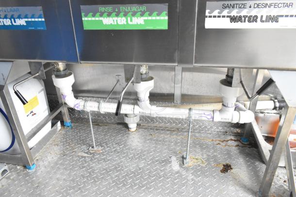 Commercial kitchen plumbing and drainage system on diamond plate flooring, labels for rinse and sanitize water lines visible.