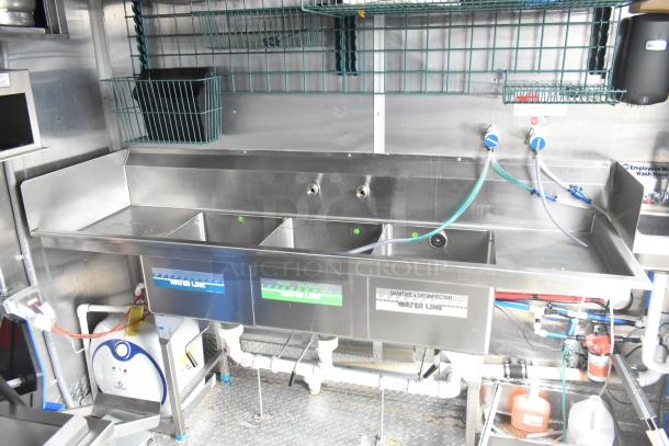 Stainless steel commercial sink with three compartments, labeled water lines, and industrial plumbing in a mobile kitchen trailer.