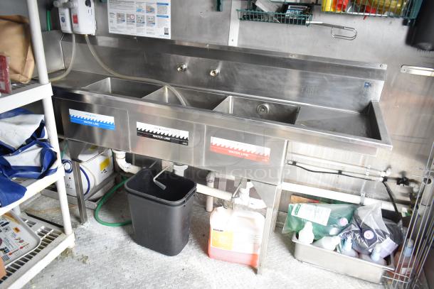 Three-compartment stainless steel sink in Rock Solid Cargo mobile kitchen, with labeled water lines. Equipped for food prep.
