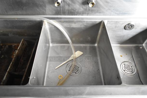 Stainless steel 3-compartment sink with drainboard in a mobile kitchen trailer, showing attached hose and scraper.