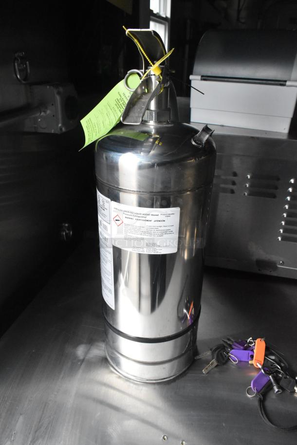 Fire suppression cylinder with label detailing contents and safety warnings; green inspection tag attached. Keys on the side.