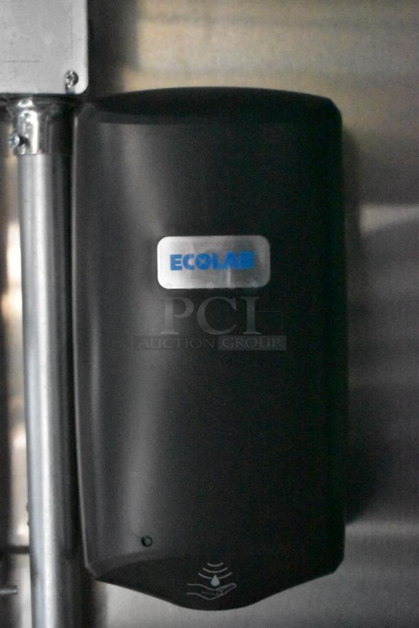 Wall-mounted Ecolab soap dispenser, black plastic, with sensor icon and Ecolab logo, set against a stainless steel surface.