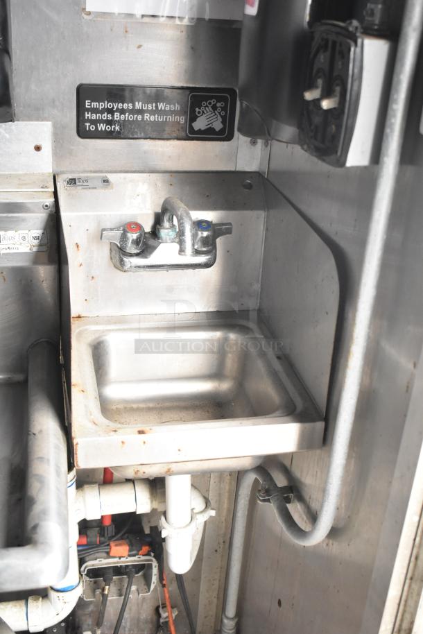 Stainless steel sink in Cruising Kitchens trailer, marked with handwashing sign. Features plumbing and faucet. Visible wear.