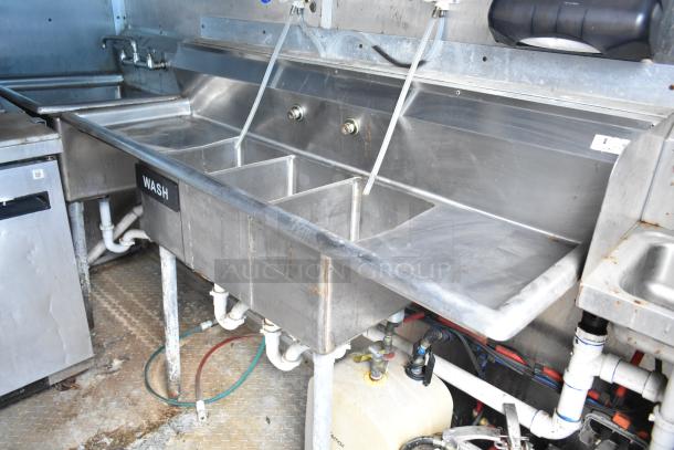 2019 Cruising Kitchens trailer interior showing stainless steel triple sink with labeled "Wash" sign, plumbing visible below.