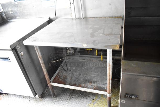 Stainless steel work table with visible wear, situated between commercial kitchen appliances, inside 2019 Cruising Kitchens trailer.