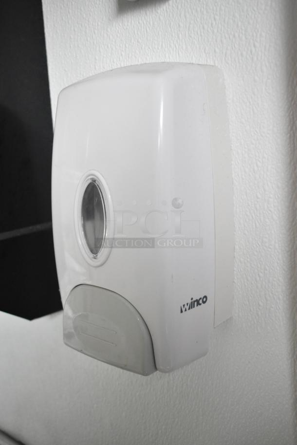 Wall-mounted Winco soap dispenser in white, attached to a textured surface.