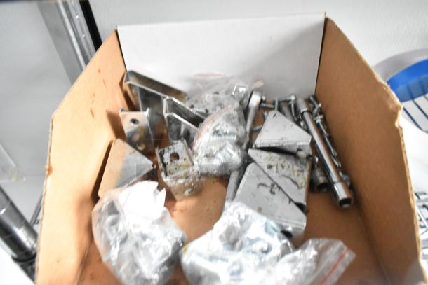 Box of metal brackets and hardware, possibly spare parts for the 2020 SDG food trailer; items are loosely organized and appear unused.
