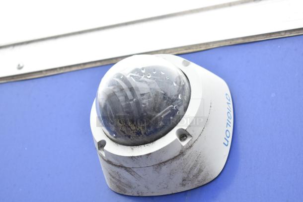 Security camera mounted on a mobile kitchen trailer, Avigilon branding visible, slightly weathered condition.