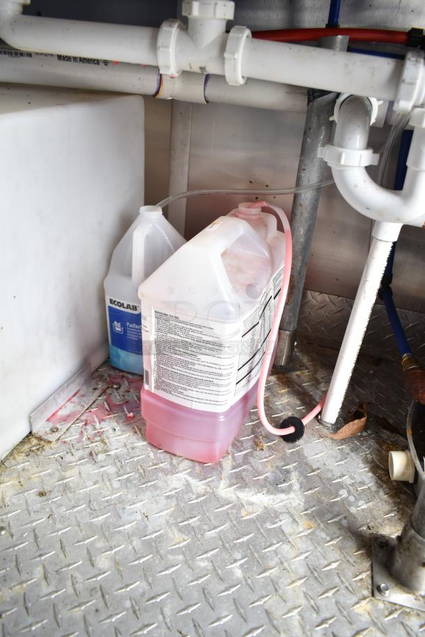 Mobile kitchen trailer interior with cleaning supplies, pipes, and metal flooring. Ecolab detergent visible in containers.