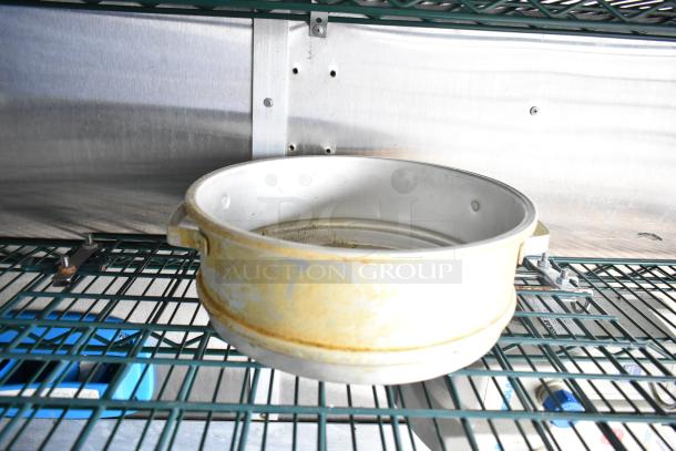 Commercial kitchen pot on a storage shelf inside a mobile kitchen trailer. Steel walls and green wire racks are visible.
